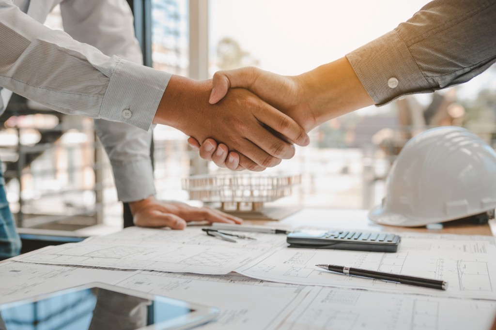 Handshake over construction drawings, a hard hat, and project tools