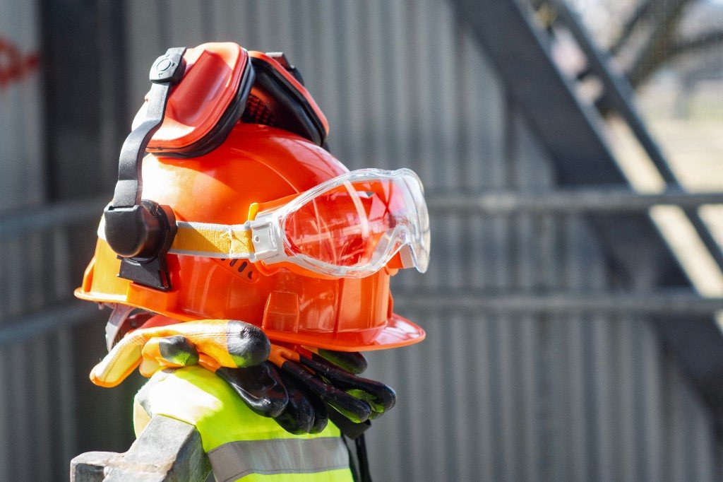 Hard hat, earmuffs, goggles, gloves, and a high-visibility vest stacked on a site railing